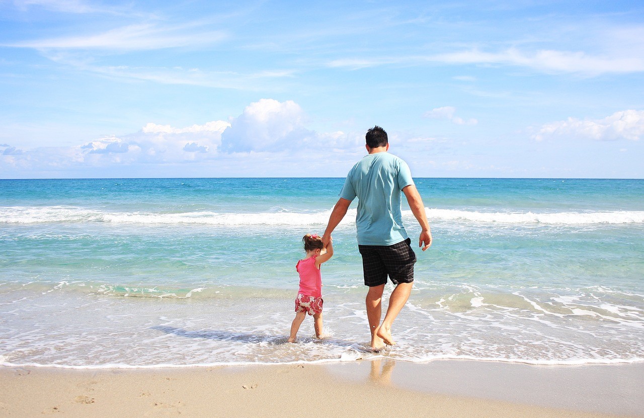 Pai andando na orla da praia de mãos dadas com filha pequena. Menina está pulando ondas enquanto pai esta despreocupado por ter trancando as portas de segurança da casa.